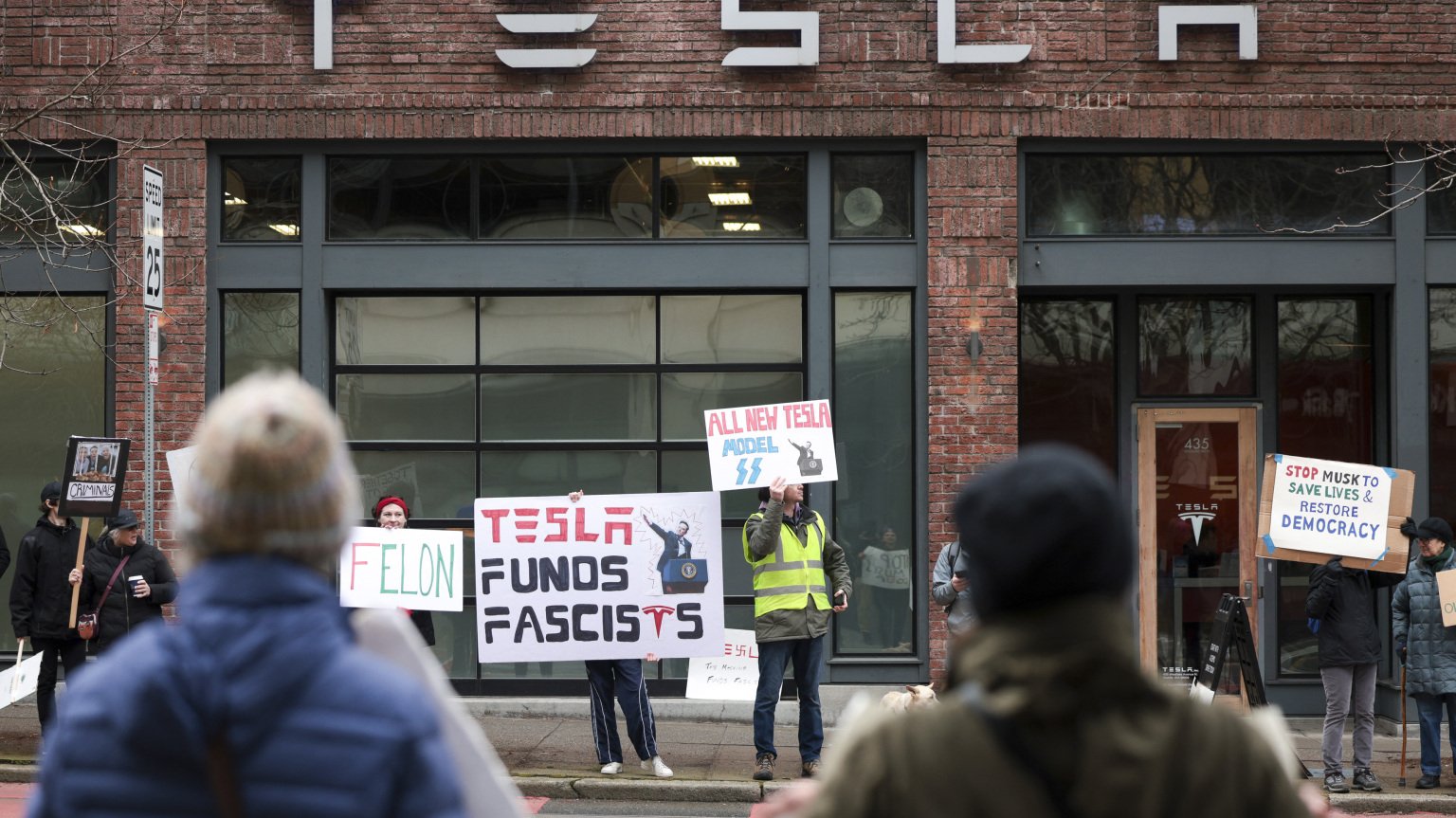 A groups stands outside a Tesla showroom holding signs, one of which says "Tesla funds fascists."