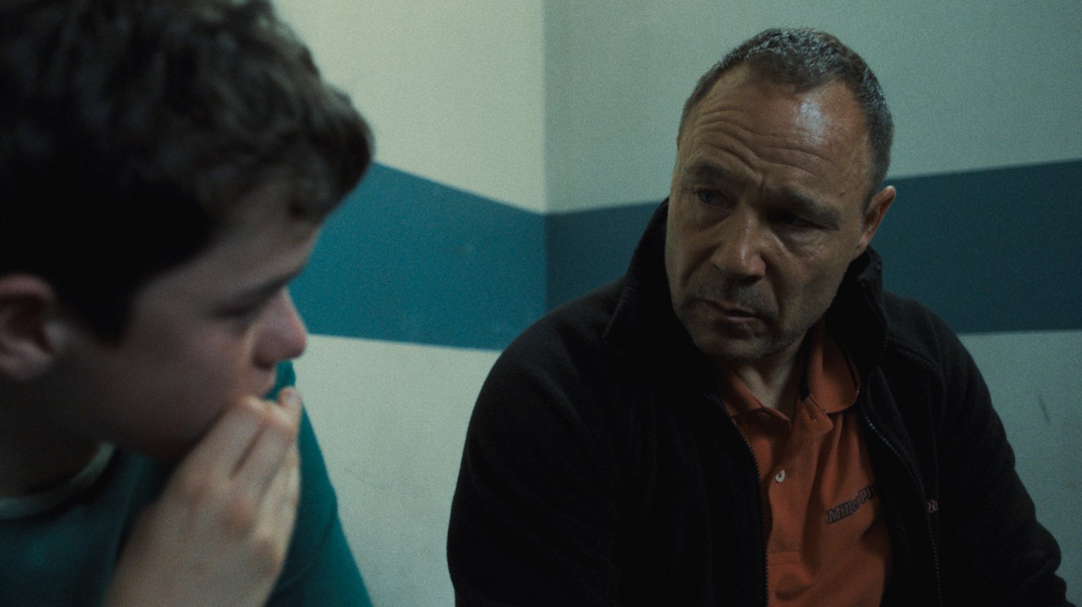 A man sits at a table in a police room looking at a teenage boy.