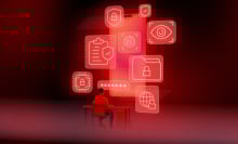 A person sits at a computer desk with numerous red screens around them. 