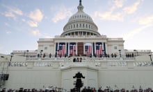 The United States Marine Band rehearsing their performance in front of the Capitol building, as large American flags drape across the facade.