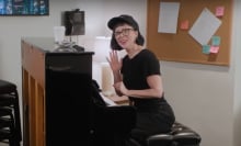 A woman sits at a piano, smiling and waving.