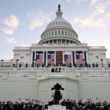 The United States Marine Band rehearsing their performance in front of the Capitol building, as large American flags drape across the facade.