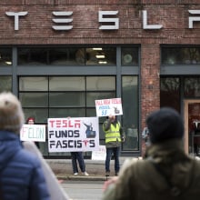 A groups stands outside a Tesla showroom holding signs, one of which says "Tesla funds fascists."