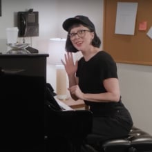 A woman sits at a piano, smiling and waving.