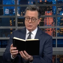 A man in a suit stands on a talk show stage reading from a black book.