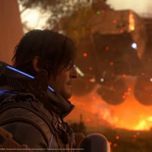 A close-up of a male character with shoulder-length dark hair and a futuristic armored suit, featuring glowing blue lights and intricate mechanical details. He gazes intently into the distance, with a determined expression. Behind him, a massive hovering machine looms over a fiery battlefield, with sparks and embers floating in the air. The scene is bathed in warm orange hues, contrasting with the cool tones of the character’s suit. The image is a high-quality, cinematic screenshot from a video game, captured on PS5, and developed by Kojima Productions.