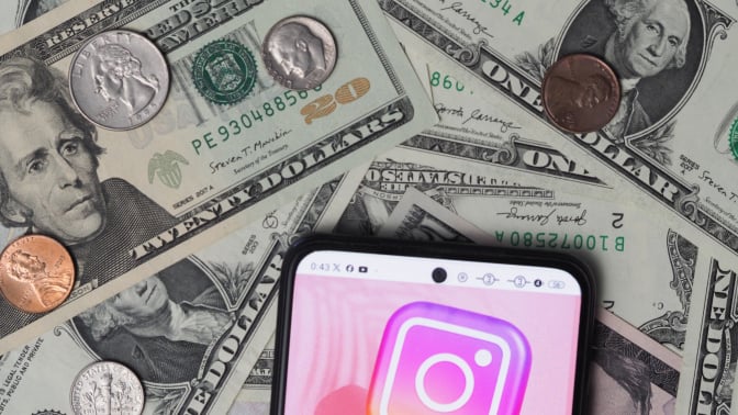 An Instagram logo seen displayed on a smartphone with United States Dollar notes and coins in the background.