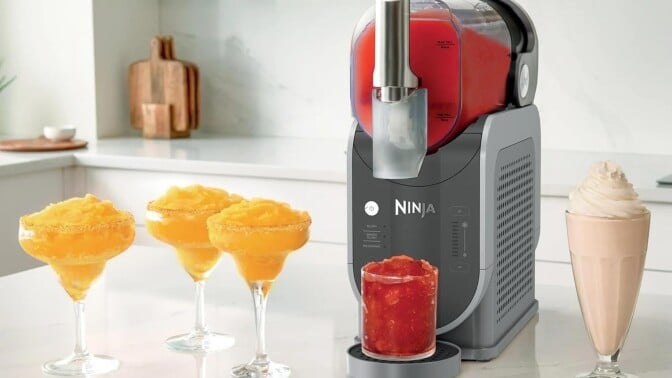 a ninja slushi machine sits on a white countertop in a kitchen with several frozen drinks surrounding the machine
