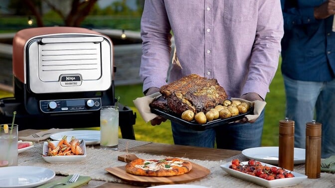 a person holds a plate of prepared food next to the ninja woodfire outdoor oven in front of a table that's filled with food