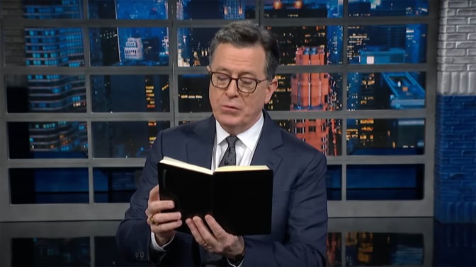 A man in a suit stands on a talk show stage reading from a black book.