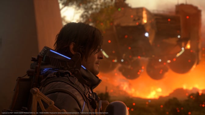 A close-up of a male character with shoulder-length dark hair and a futuristic armored suit, featuring glowing blue lights and intricate mechanical details. He gazes intently into the distance, with a determined expression. Behind him, a massive hovering machine looms over a fiery battlefield, with sparks and embers floating in the air. The scene is bathed in warm orange hues, contrasting with the cool tones of the character’s suit. The image is a high-quality, cinematic screenshot from a video game, captured on PS5, and developed by Kojima Productions.