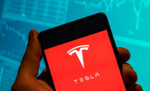 The Tesla logo is seen displayed on a smartphone with an economic stock exchange index graph in the background.