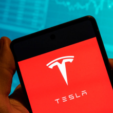 The Tesla logo is seen displayed on a smartphone with an economic stock exchange index graph in the background.